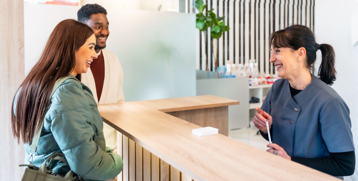 Receptionist greeting two smiling visitors at desk.